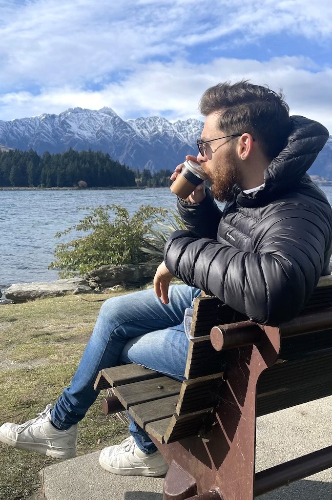 Paul drinking coffee beside a lake and mountains.